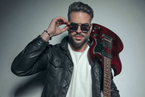 pohledný rockový kytarista drží elektrickou kytaru na rameni a aranžuje si sluneční brýle  - Fotografie, Obrázek