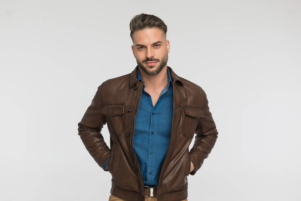 sexy unshaved man holding hands in brown leather jacket pockets and being confident in front of light grey background in studio - Foto, Imagen