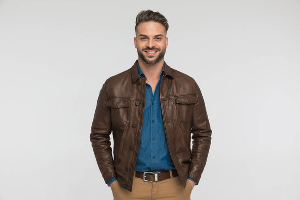 gelukkig man dragen bruin lederen jas houden handen in zakken en glimlachen terwijl poseren op grijze achtergrond in de studio - Foto, afbeelding