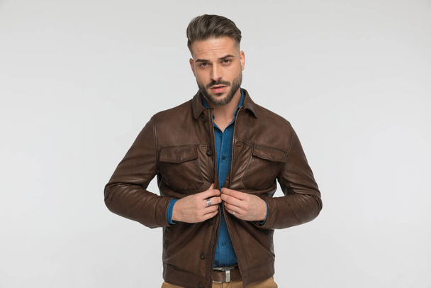 fashion man with beard closing brown leather jacket and being confident on grey background in studio - Photo, Image
