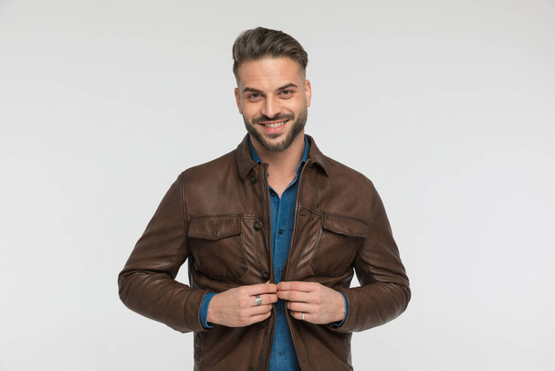 homem casual animado com camisa jeans abotoando jaqueta de couro marrom e sorrindo no fundo cinza - Foto, Imagem