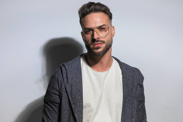 guapo barbudo con gafas con capucha y posando de una manera seria delante de fondo gris claro en el estudio - Foto, imagen
