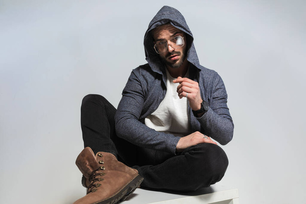 handsome man with hoodie on his head crossing legs in a fashion pose while holding arms and posing in front of grey background - Photo, Image