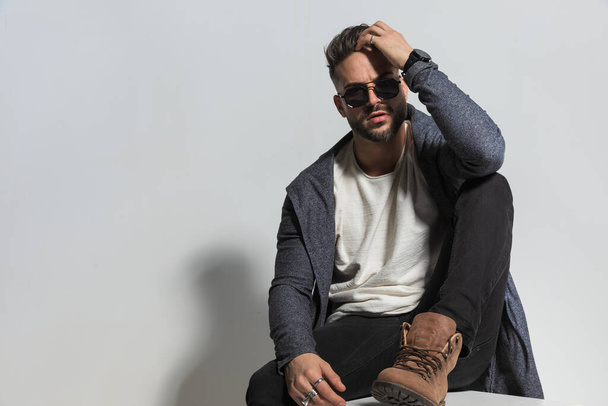 picture of sexy casual man with sunglasses adjusting hairstyle and being cool while holding elbow on knee and posing with leg up on grey background in studio - Valokuva, kuva