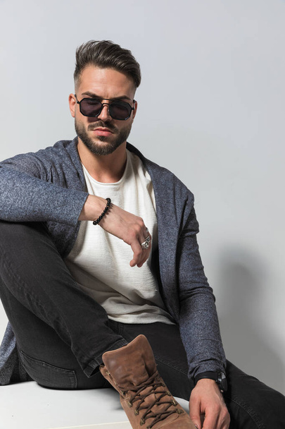 cutout picture of bearded stylish guy in hoodie with sunglasses holding leg up and posing with elbow on knee in front of grey background in studio - Photo, Image