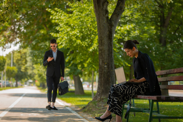 A gorgeous businesswoman is working outdoors, sitting on a bench while a handsome male businessman is passing in front of her - Foto, Imagen