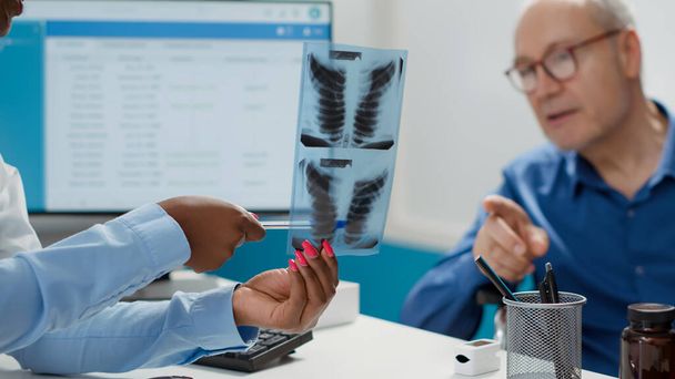 General practitioner analyzing x ray scan with senior man at checkup visit appointment. Doctor explaining radiography results and bones diagnosis to cure patient with physical impairment. - Foto, imagen