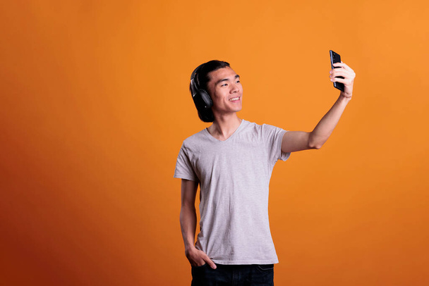 スマートフォンのフロントカメラで自撮りをするワイヤレスヘッドフォンでアジアのティーンエイジャーを笑顔。携帯電話を持っているイヤホンで若い陽気な魅力的な男,写真のポーズ - 写真・画像