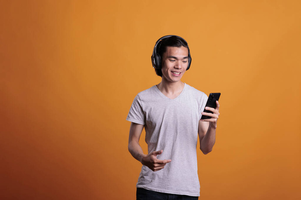 Man in wireless headphones watching video on mobile phone, smiling asian teenager listening to podcast. Young cheerful person wearing earphones, enjoying music on smartphone - 写真・画像