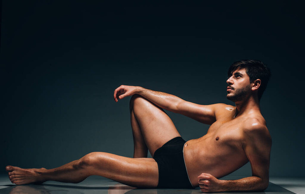Sexy portrait of a very muscular shirtless male model in underwear against black wall in sensual pose - Фото, изображение