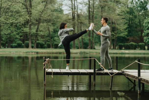 Niesamowita dziewczyna wykonuje ruch nogą, a jej chłopak pomaga jej, gdy stoją na moście w parku. - Zdjęcie, obraz