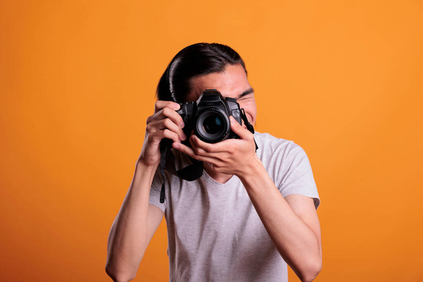 Fotógrafo tomando fotos en cámara dslr profesional, joven hombre asiático fotografiando, vista frontal plano medio. Adolescente mirando en visor de dispositivos, hobby creativo, sesión de fotos - Foto, imagen