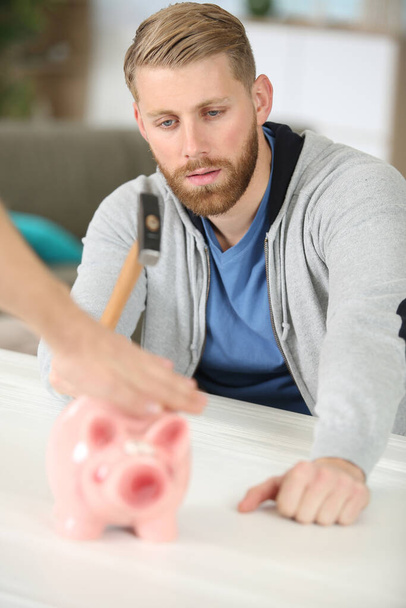 young man breaking piggy bank with hammer at table indoors - Fotoğraf, Görsel