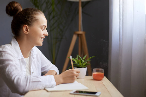 Profile portrait of smiling beautiful woman writing notes in paper notebook, thinking about something pleasant, dreaming, sitting at the table by the window. - Foto, Imagen