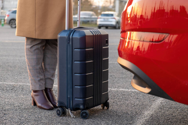 A woman in a brown coat stands with a suitcase near a red car. Shuttle from an airport concept  - Фото, изображение