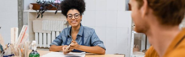 happy african american woman in eyeglasses molding and looking at redhead man on blurred foreground, banner - Фото, изображение