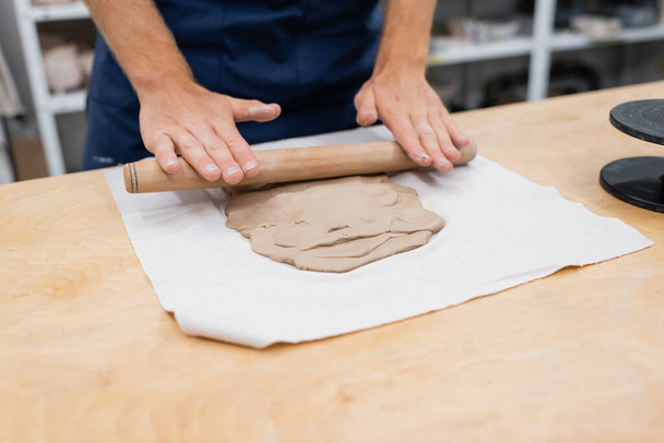 vista recortada del hombre en delantal que forma la pieza de arcilla con rodillo durante la clase de cerámica  - Foto, imagen
