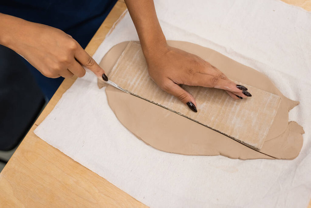 bovenaanzicht van afrikaanse Amerikaanse vrouw snijden klei met mes in de buurt van karton  - Foto, afbeelding