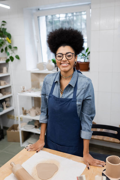 happy young african american woman in eyeglasses and apron standing near rolling pin in pottery workshop - Foto, Bild
