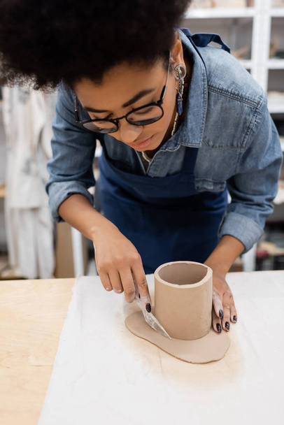 ricci donna africana americana in occhiali da vista taglio argilla intorno tazza sagomata con coltello durante la classe di ceramica - Foto, immagini