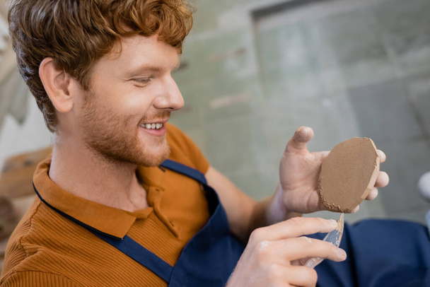 cheerful and redhead man in apron cutting piece of clay with knife - Foto, afbeelding