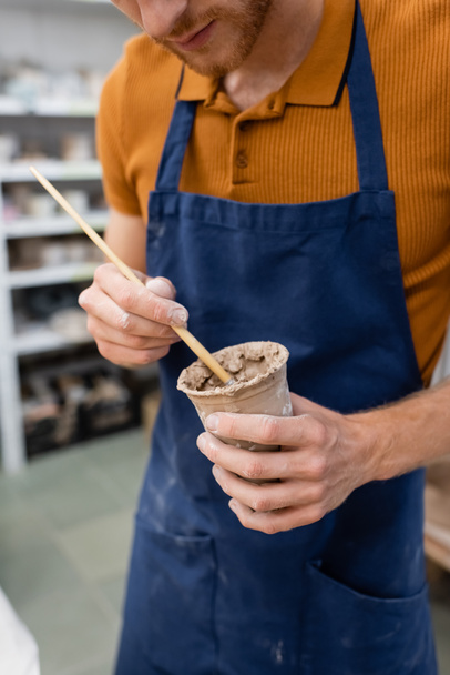 cropped view of bearded man holding paintbrush and clay cup in hands - Foto, Bild