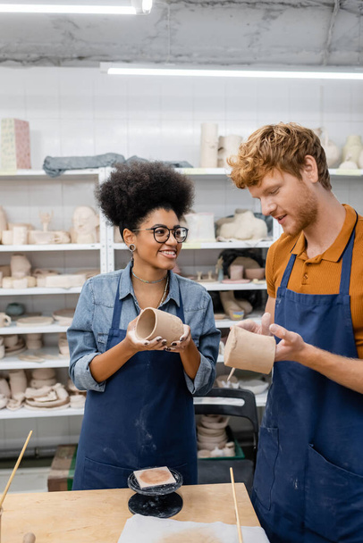 cheerful interracial couple molding clay cups during date in pottery studio  - Foto, afbeelding