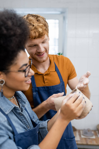 happy redhead man pointing with hand at clay cup during date with african american girlfriend in pottery studio  - Photo, Image