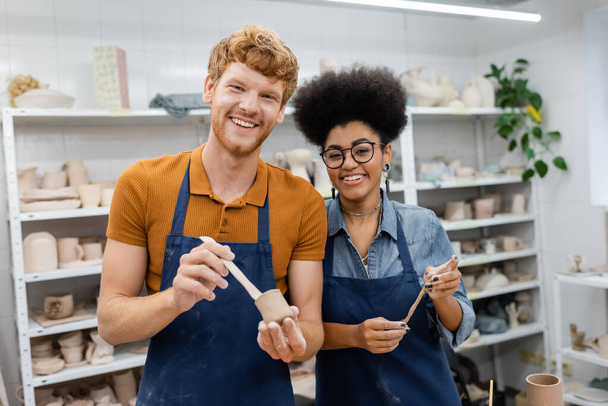 Улыбающийся мужчина и африканская американка держат глину в керамической мастерской  - Фото, изображение