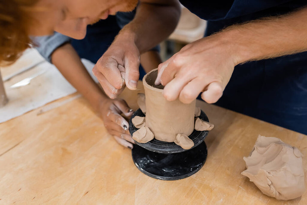 Hombre borroso formando taza de arcilla cerca de profesor afroamericano en taller de cerámica  - Foto, imagen