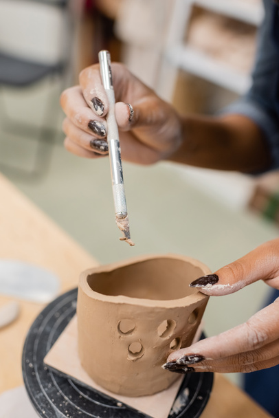 Vista ritagliata dell'artigiana afro-americana che tiene lo strumento vicino al prodotto in argilla in officina  - Foto, immagini