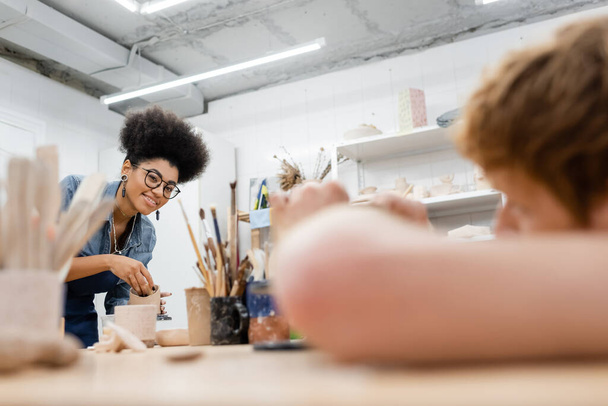 Sonriente mujer afroamericana sosteniendo arcilla cerca de herramientas borrosas y novio en taller de arcilla  - Foto, imagen