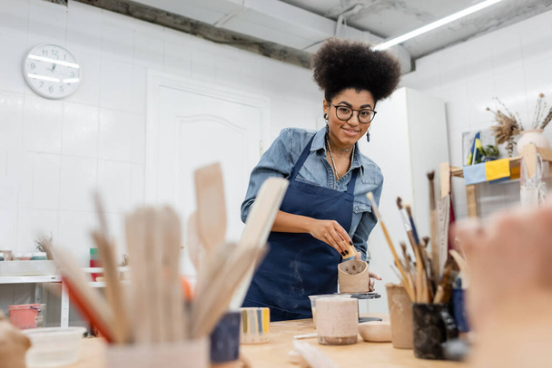 Lächelnder afrikanisch-amerikanischer Kunsthandwerker in Brille mit Schwamm und Ton, während er in Töpferwerkstatt in die Kamera blickt  - Foto, Bild