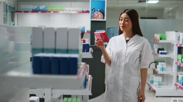 Веселий азіатський працівник вивчає фармацевтичні продукти з полиць аптек, дивлячись на аптечки і таблетки пляшок. Жіночий фармацевт перевіряє вітаміни та наркотики
. - Кадри, відео