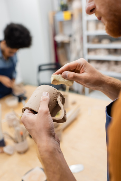 Cropped view of man holding clay cup and sponge during date with blurred african american girlfriend in pottery workshop  - Photo, Image