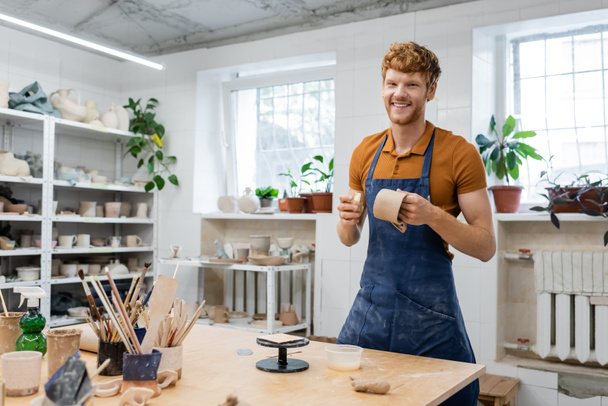 Uśmiechnięty, rudy rzemieślnik w fartuchu patrzący w kamerę podczas wykonywania ceramicznych filiżanek w warsztacie  - Zdjęcie, obraz