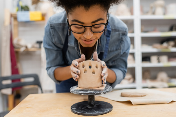 Sorridente donna afroamericana in occhiali che forma scultura in argilla in studio di ceramica  - Foto, immagini