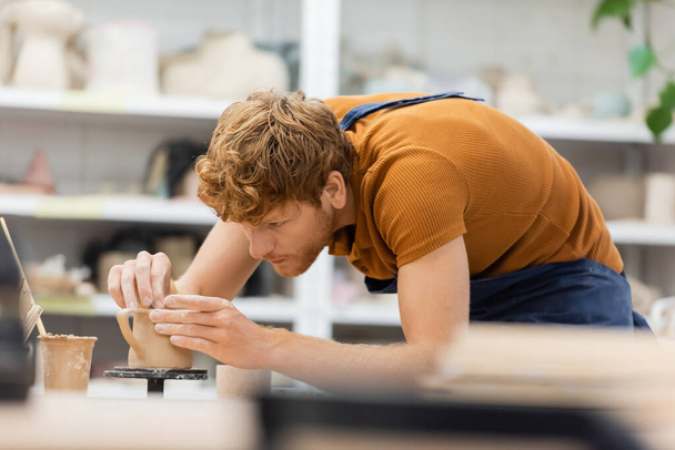 Redhead master v zástěře vytváří hliněný pohár v keramické dílně  - Fotografie, Obrázek