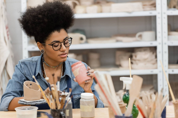 Artesana afroamericana en anteojos sosteniendo tarro con pintura cerca de pinceles borrosos y escultura de arcilla en estudio de cerámica  - Foto, Imagen
