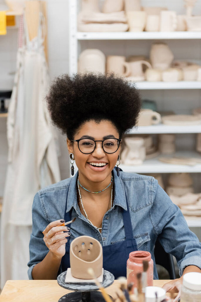glimlachende Afrikaans-Amerikaanse vrouw in bril met penseel in de buurt van klei beeldhouwkunst en verf in aardewerk workshop  - Foto, afbeelding