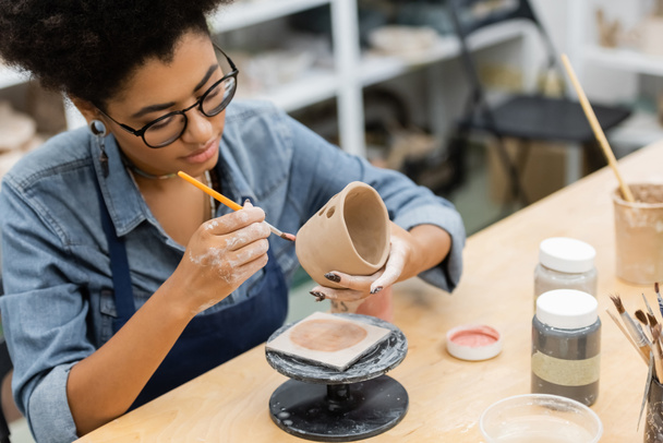 Artigiano afroamericano in grembiule dipinto su prodotto ceramico vicino a vasi con vernice in studio di ceramica  - Foto, immagini