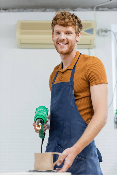 Lächelnder rothaariger Handwerker in Schürze mit Wärmepistole in der Nähe verschwommener Keramiktasse in Töpferwerkstatt  - Foto, Bild