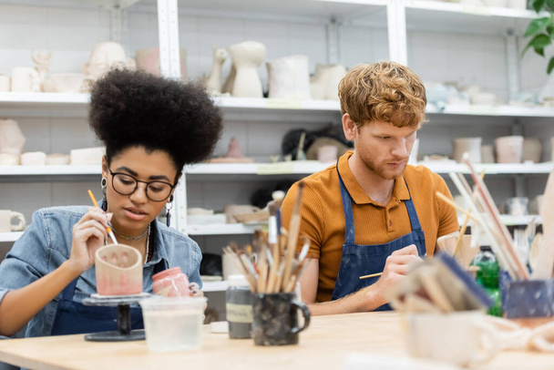 Interracial couple in aprons painting on clay sculptures during date in pottery studio  - Fotoğraf, Görsel