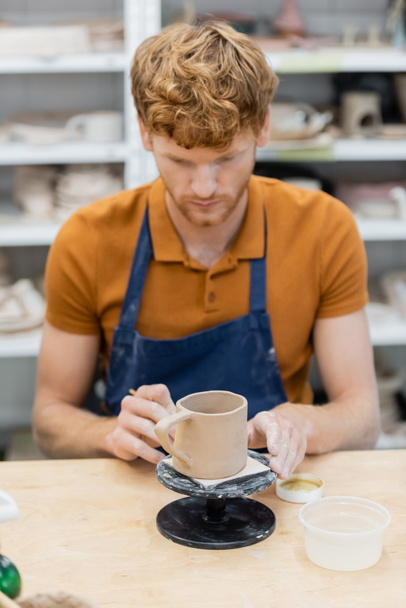 Pelirroja artesana en delantal mirando taza de cerámica en taller de cerámica  - Foto, Imagen