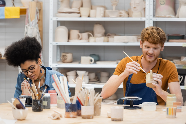 Positive multiethnic couple painting clay sculptures during date in pottery studio  - Photo, Image
