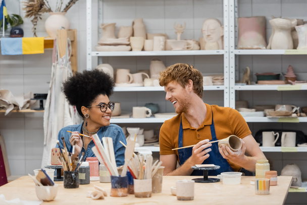 Homme souriant regardant petite amie afro-américaine près de l'argile et pinceaux en atelier de poterie  - Photo, image