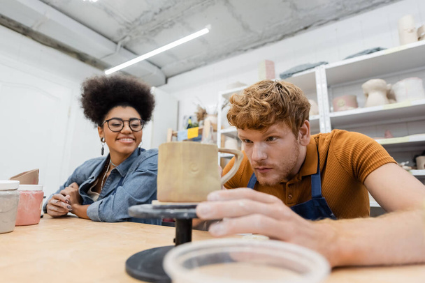 Redhead man looking at clay cup near smiling african american girlfriend in pottery studio  - Photo, Image