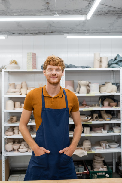 Smiling redhead artisan in apron looking at camera in pottery studio  - Φωτογραφία, εικόνα