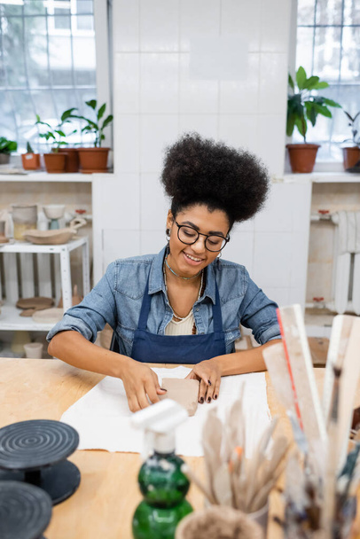 Mujer afroamericana sonriente en delantal sosteniendo arcilla en la mesa en taller de cerámica  - Foto, Imagen