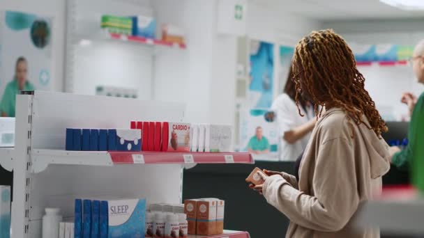 Cliente afroamericano feliz marcando cajas de medicamentos en los estantes de farmacia, mirando productos de salud para comprar tratamiento con receta. Mujer comprando medicamentos y vitaminas. - Metraje, vídeo